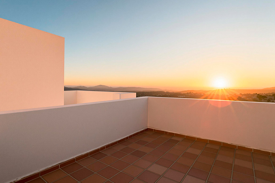 A modern rooftop terrace with clean white walls and warm terracotta floor tiles. The open space overlooks distant hills and a wide landscape, giving a minimal and airy Mediterranean-inspired feel. The geometric lines of the architecture create a simple, elegant, and uncluttered environment.