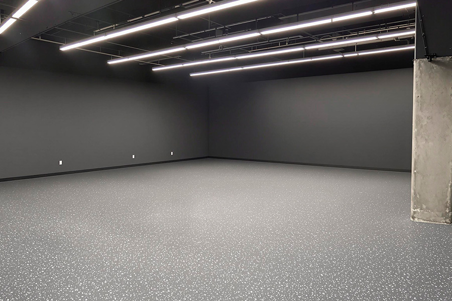 A spacious industrial-style studio with matte charcoal walls and a speckled gray epoxy floor. The ceiling features exposed structural elements and multiple long LED light bars arranged in parallel rows. A concrete pillar stands on the right side, adding a raw, utilitarian touch. The space feels modern, minimal, and functional.
